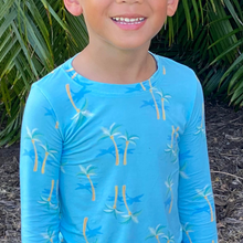Load image into Gallery viewer, A toddler wearing a long-sleeve blue pajama top with a palm tree print and matching pants, standing outdoors.