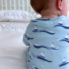 Load image into Gallery viewer, A baby wearing a blue zippered romper with a sailboat and seagull print, sitting on a bed with a white bedding.