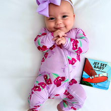 Load image into Gallery viewer, A baby wearing a long-sleeved purple floral zippered romper with a large bow on the head, sitting next to a book titled 'Jet Set Baby'.