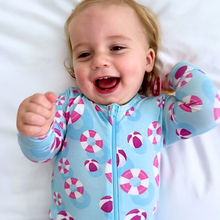 Load image into Gallery viewer, A baby wearing a blue zippered romper with pink and purple pool float patterns, smiling and lying down.
