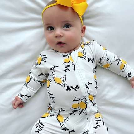 A baby wearing a white zippered romper with a pattern of yellow lemons and black leaves, accompanied by a yellow headband.