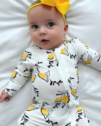 A baby wearing a white zippered romper with a pattern of yellow lemons and black leaves, accompanied by a yellow headband.