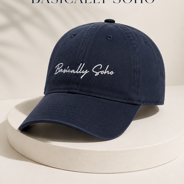 A navy blue baseball cap with 'Basically Soho' embroidered in white script on the front, displayed on a white pedestal with promotional text overlays.