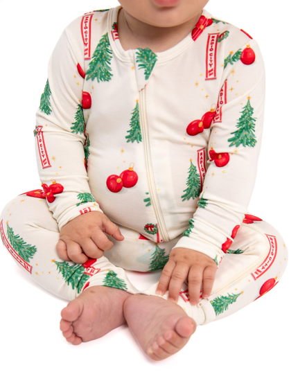 A baby wearing a cream-colored zip-up one-piece pajama with a festive Christmas print of green trees, red ornaments, and red banners, sitting cross-legged against a white background.
