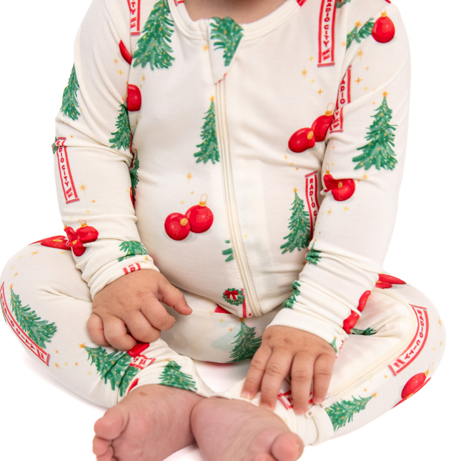 A baby wearing a cream-colored zip-up one-piece pajama with a festive Christmas print of green trees, red ornaments, and red banners, sitting cross-legged against a white background.