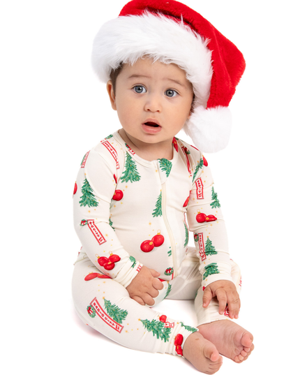 A baby wearing a festive Christmas-themed footed sleeper and a red Santa hat, sitting against a solid white background.