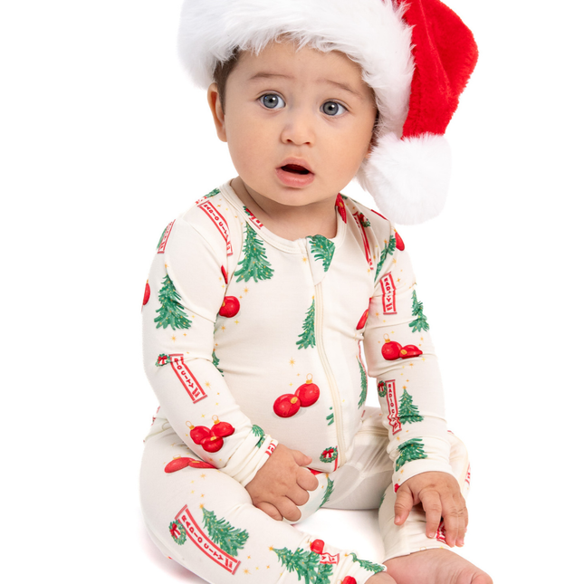 A baby wearing a festive Christmas-themed footed sleeper and a red Santa hat, sitting against a solid white background.