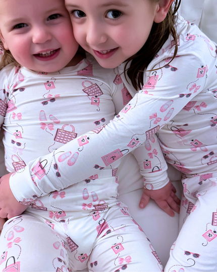 Two toddlers wearing matching pink pajamas with various designs, including purses and ballet slippers, sitting close together and smiling.