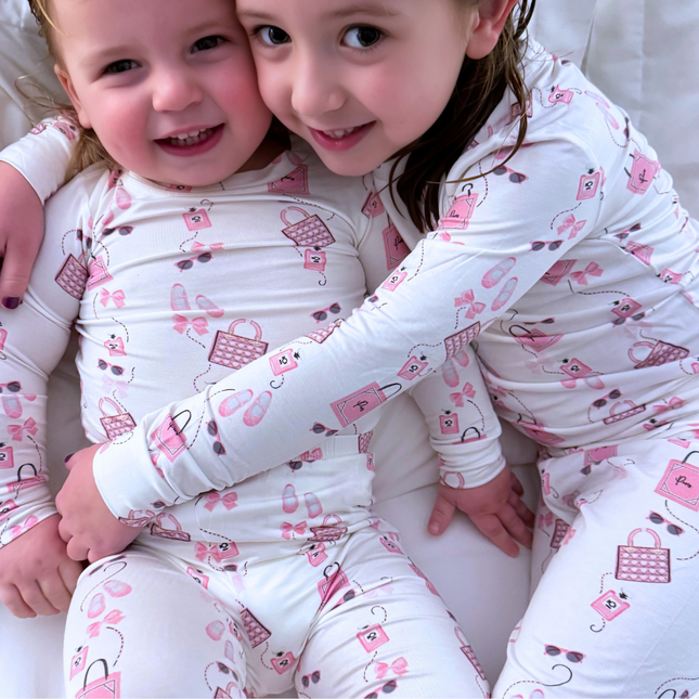 Two toddlers wearing matching pink pajamas with various designs, including purses and ballet slippers, sitting close together and smiling.