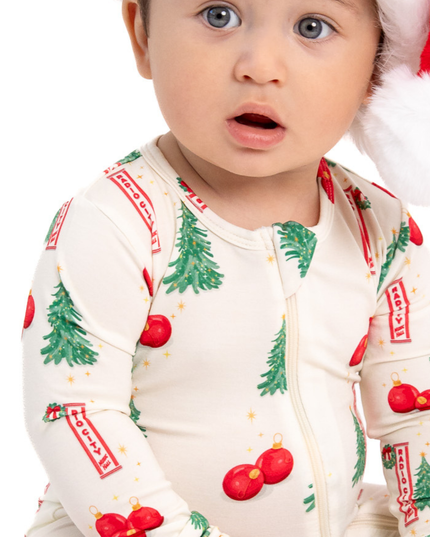 A close-up of a baby wearing a cream-colored Christmas-themed long-sleeve zip-up sleeper with green pine trees, red ornaments, and red-and-white striped banners, along with a Santa hat.