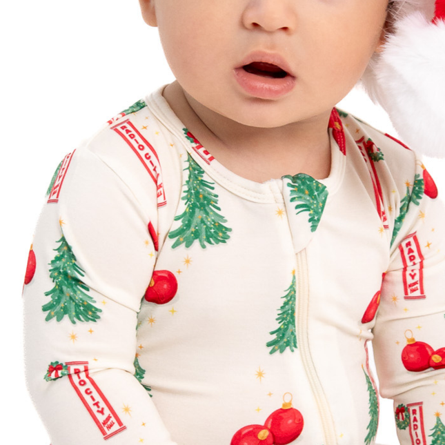 A close-up of a baby wearing a cream-colored Christmas-themed long-sleeve zip-up sleeper with green pine trees, red ornaments, and red-and-white striped banners, along with a Santa hat.