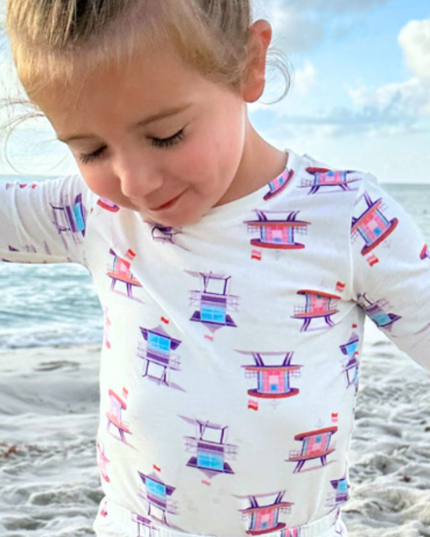 A toddler wearing a pink two-piece pajama set with a print of lifeguard towers on it, standing on a beach.