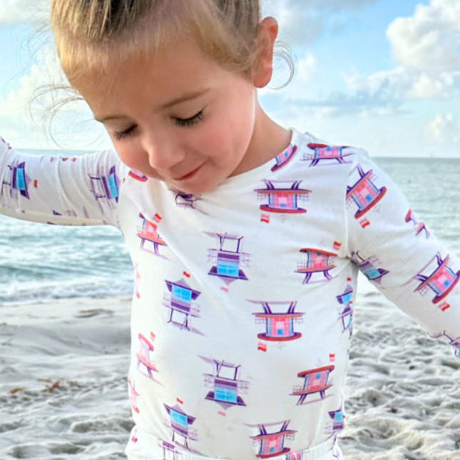 A toddler wearing a pink two-piece pajama set with a print of lifeguard towers on it, standing on a beach.