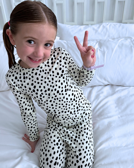 Young girl in a polka dot outfit making a peace sign on a white bed.