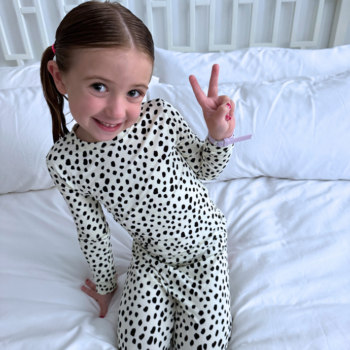 Young girl in a polka dot outfit making a peace sign on a white bed.