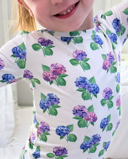 A toddler wearing a white two-piece pajama set with a floral hydrangea pattern in pink, purple, and blue.