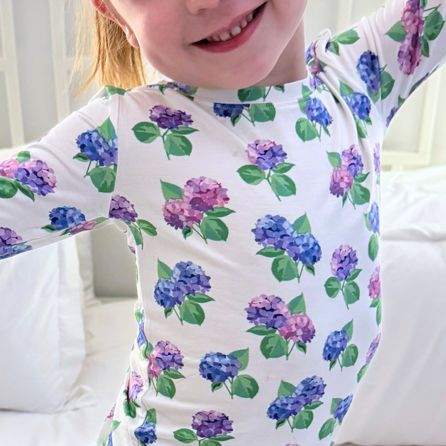 A toddler wearing a white two-piece pajama set with a floral hydrangea pattern in pink, purple, and blue.
