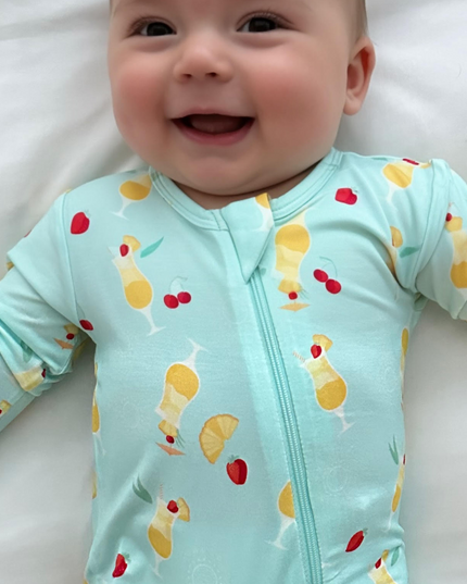 A baby wearing a light blue zippered romper with a fruity print, lying on a white surface.