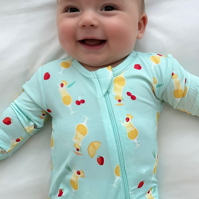 A baby wearing a light blue zippered romper with a fruity print, lying on a white surface.