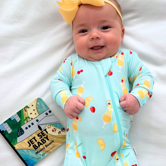 Baby in a colorful onesie with a yellow bow, lying on a white blanket next to a book.
