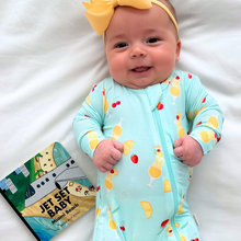 Load image into Gallery viewer, Baby in a colorful onesie with a yellow bow, lying on a white blanket next to a book.
