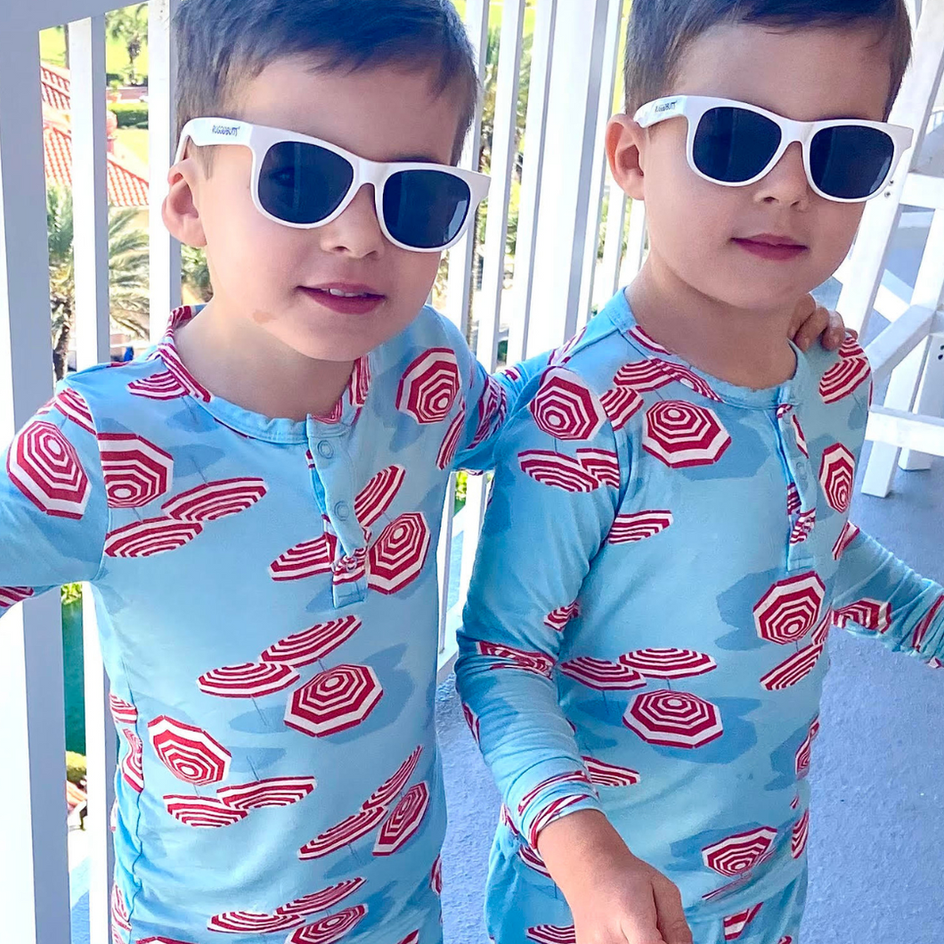 Two children wearing matching blue swimsuits with red umbrella pattern and sunglasses outdoors.