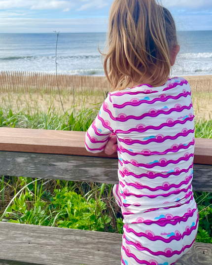 A toddler in a striped two-piece sleepwear set with a pink and white pattern, sitting on a wooden fence looking towards the ocean.