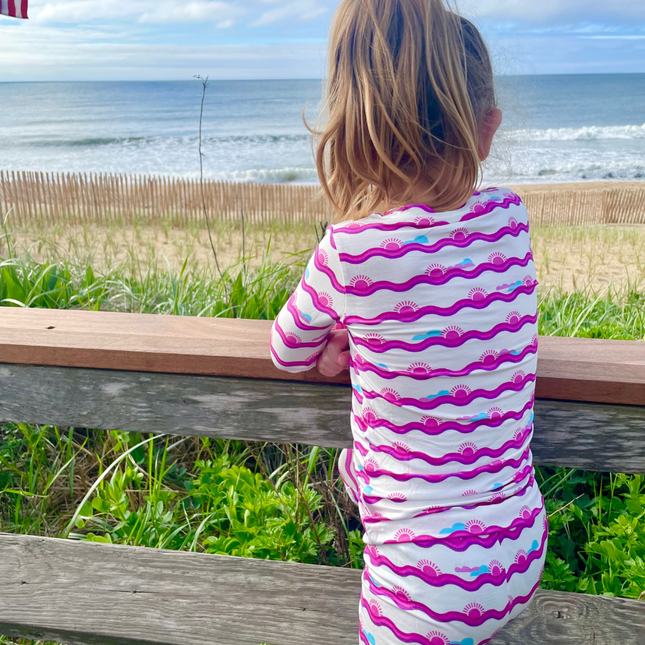 A toddler in a striped two-piece sleepwear set with a pink and white pattern, sitting on a wooden fence looking towards the ocean.