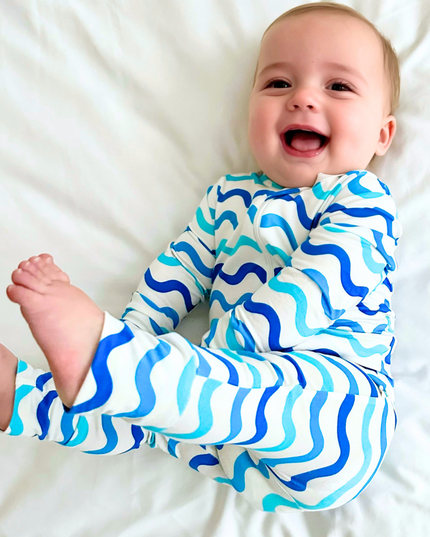 A smiling baby lying on a white bedsheet, wearing a long-sleeved footless zippered romper with a wave print in shades of blue.