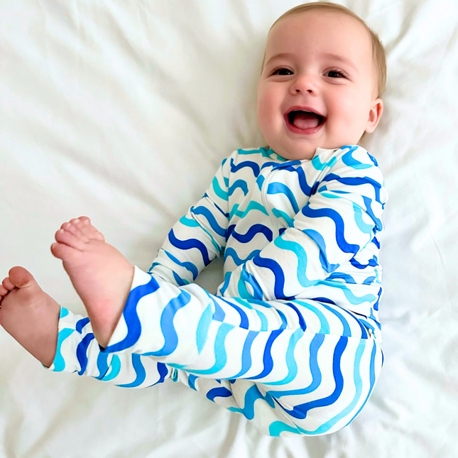 A smiling baby lying on a white bedsheet, wearing a long-sleeved footless zippered romper with a wave print in shades of blue.