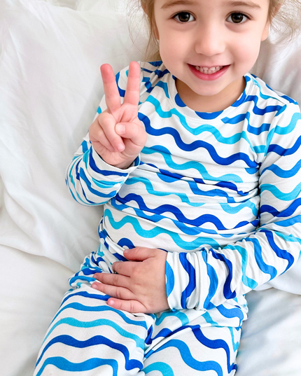 A toddler in a happy mood, sitting on a bed, wearing a two-piece pajama set with a wave print in shades of blue on a white background.