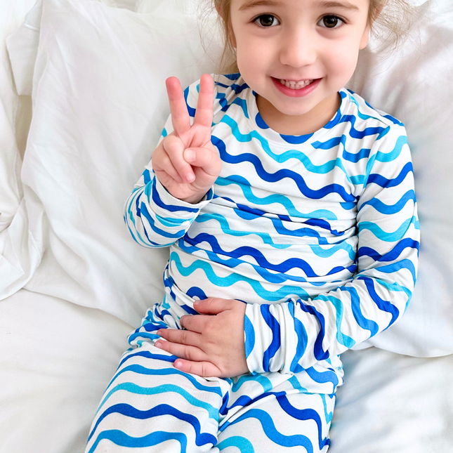A toddler in a happy mood, sitting on a bed, wearing a two-piece pajama set with a wave print in shades of blue on a white background.